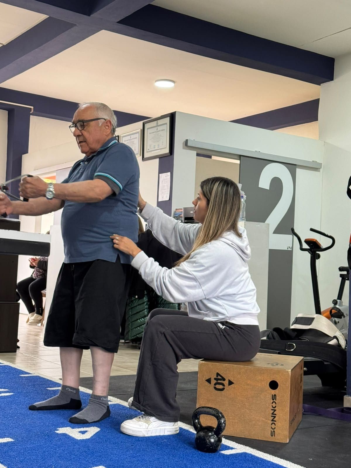 Un adulto mayor trabajando con banda elástica sobre la pista del gimnasio, acompañado por una kinesióloga de PhysioMove que le corrige la postura