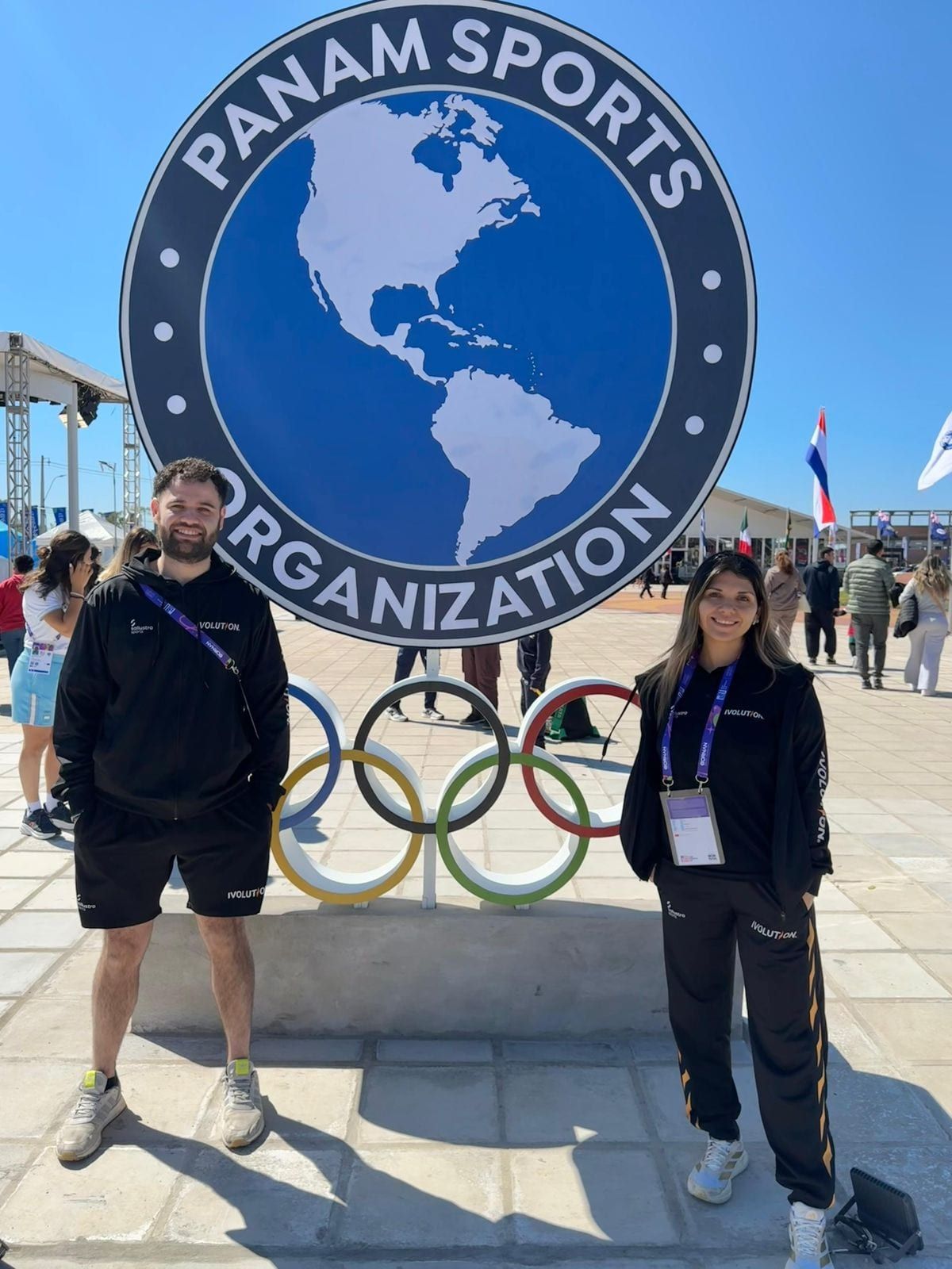 Dos profesionales de PhysioMove frente al cartel de Panam Sports con los anillos olímpicos