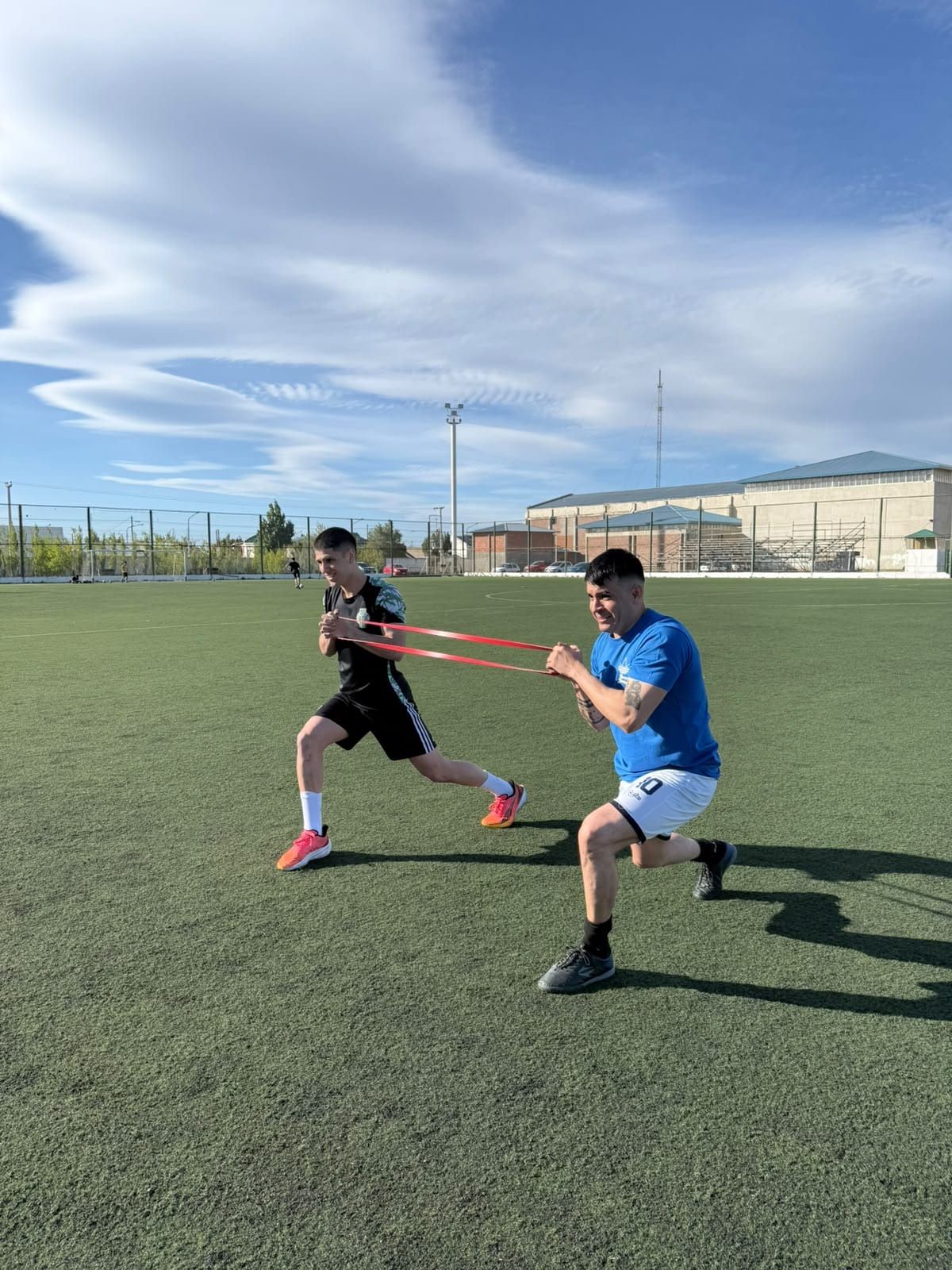 Dos personas entrenando con banda elástica en una cancha de césped sintético: una avanza contra la resistencia y la otra la sostiene